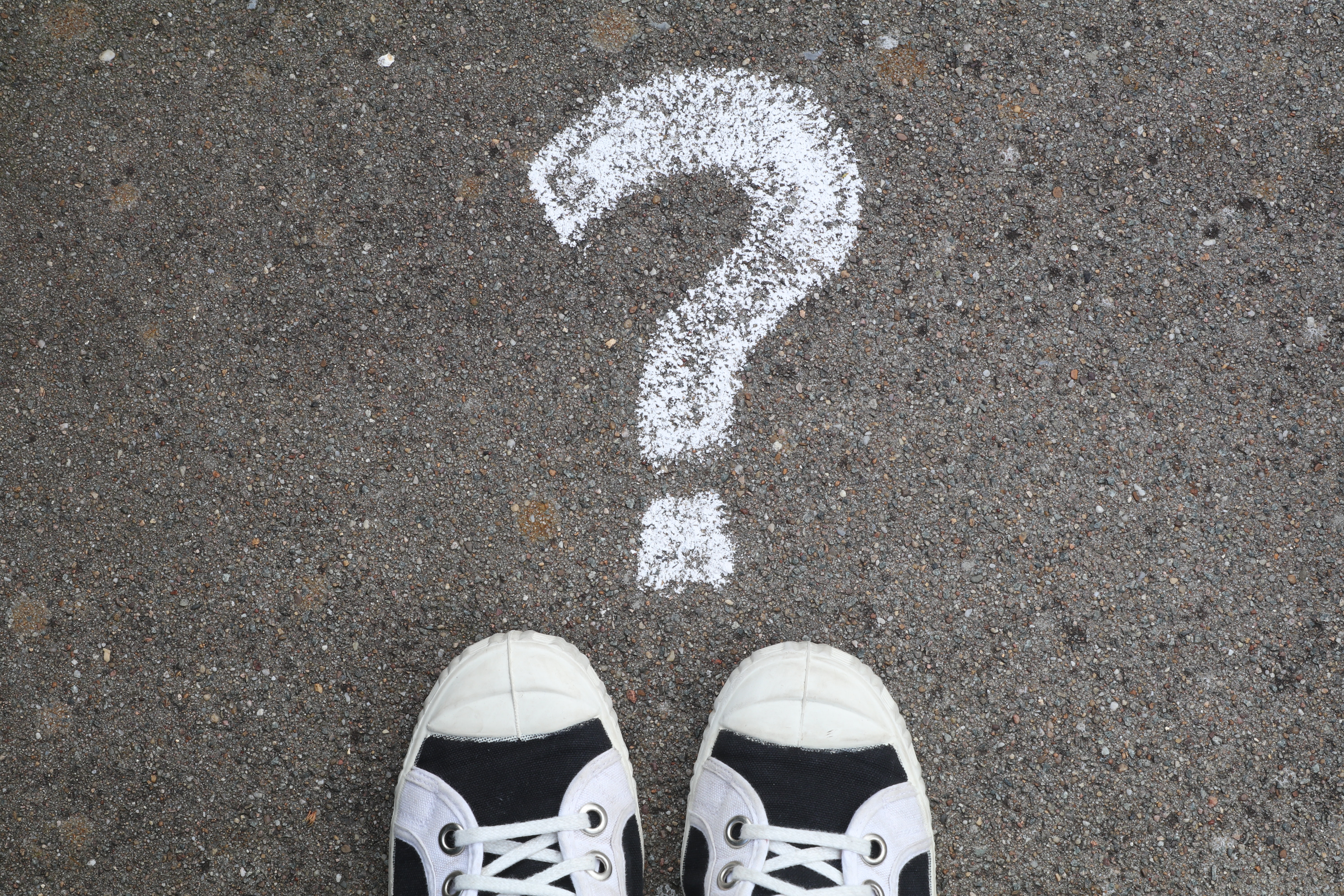 A white chalk question mark on the pavement right above the toes of two sneakers